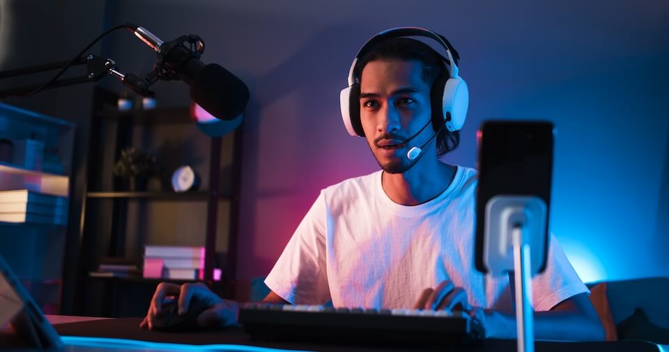 A person wearing a headset sits at a desk with a microphone, using a keyboard and computer, in a dimly lit room.