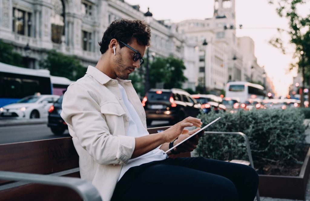 Man wearing glasses and earphones uses a tablet while sitting on a bench in a busy urban street setting.