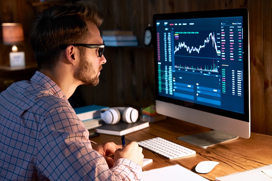 A person wearing glasses studies financial graphs on a computer screen, with a keyboard, mouse, and headphones nearby.