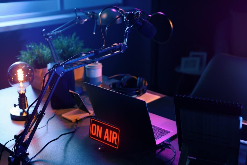 Podcast recording setup with microphone, laptop, headphones, and illuminated "ON AIR" sign on a desk in dim lighting.