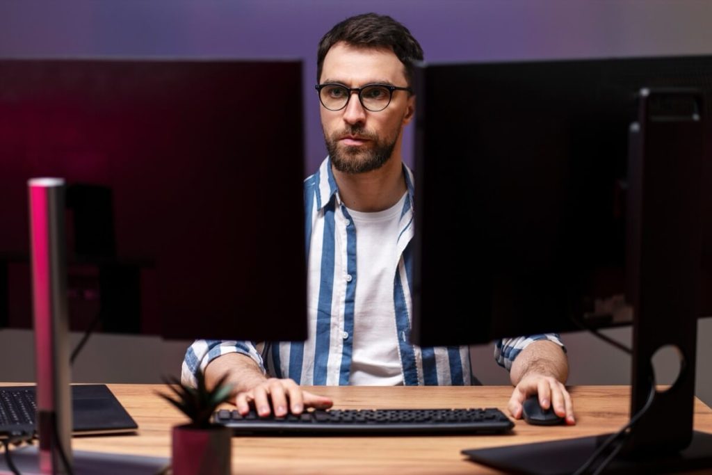 Man wearing glasses and striped shirt working at a desk with two computer monitors, focused on the screens.