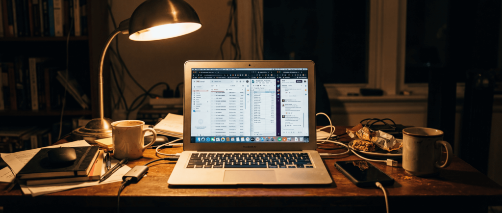 Laptop on a cluttered desk with open browser tabs, surrounded by books, mugs, a lamp, and snacks.