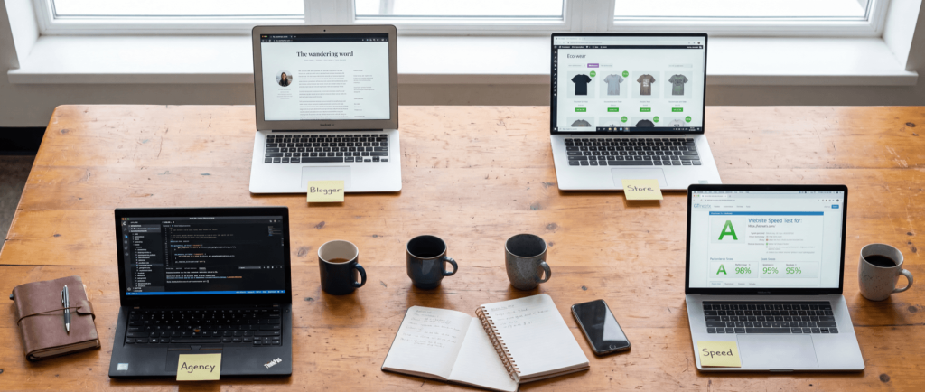 Four laptops on a wooden table display different websites, labeled "Agency," "Blogger," "Store," and "Speed," with notebooks and mugs.