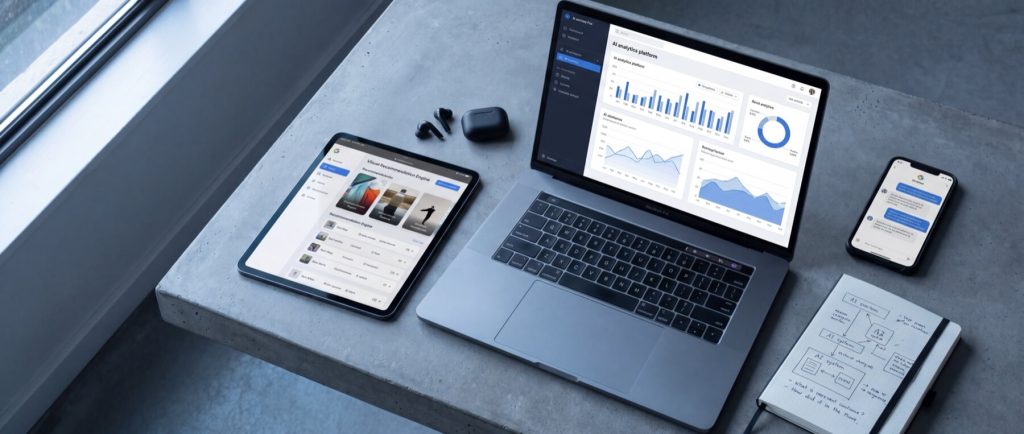 Laptop, tablet, and smartphone displaying analytics and communication apps on a concrete desk, alongside a notebook with sketches.