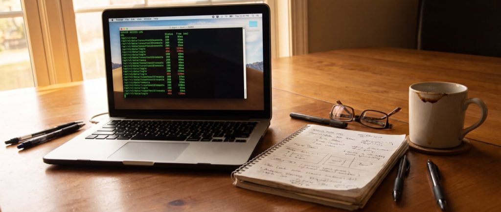 Laptop displaying server access log, notebook with handwritten notes, glasses, pens, and a coffee mug on a wooden table.
