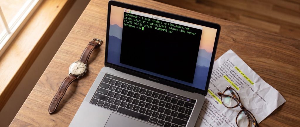 Open laptop displaying terminal commands on a wooden desk, next to a wristwatch, glasses, and a printed document.