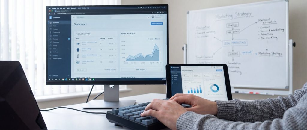 Person typing on a keyboard, viewing analytics dashboard on monitor and tablet, with marketing strategy whiteboard in background.