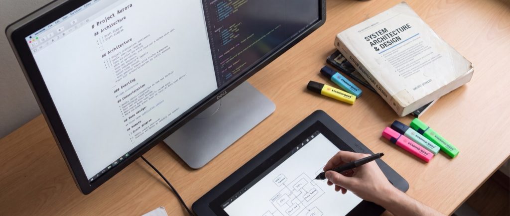 Computer monitor displaying "Project Aurora" document, tablet with flowchart, hand drawing, book titled "System Architecture & Design," highlighters.